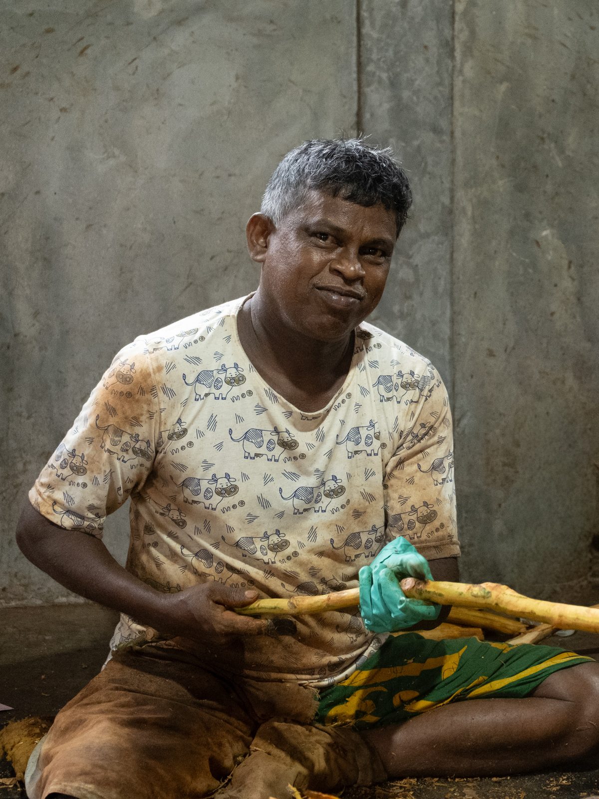 A man sitting on a concrete floor peeling a cinnamon stem, smiling at the camera
