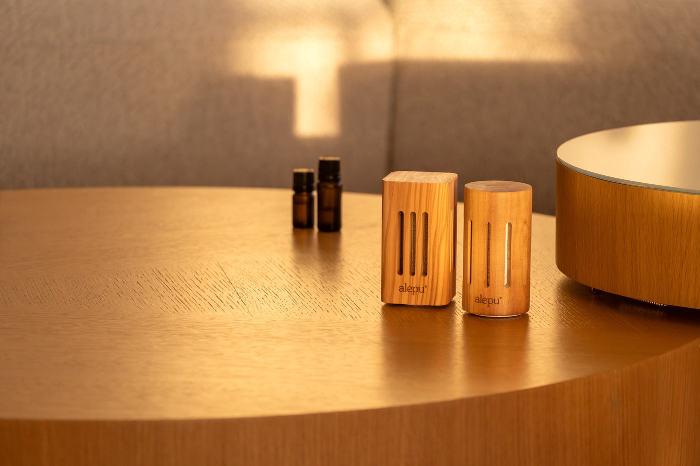 The square and round olive wood diffusers beside two bottles of oil on a round wooden table in warm evening light