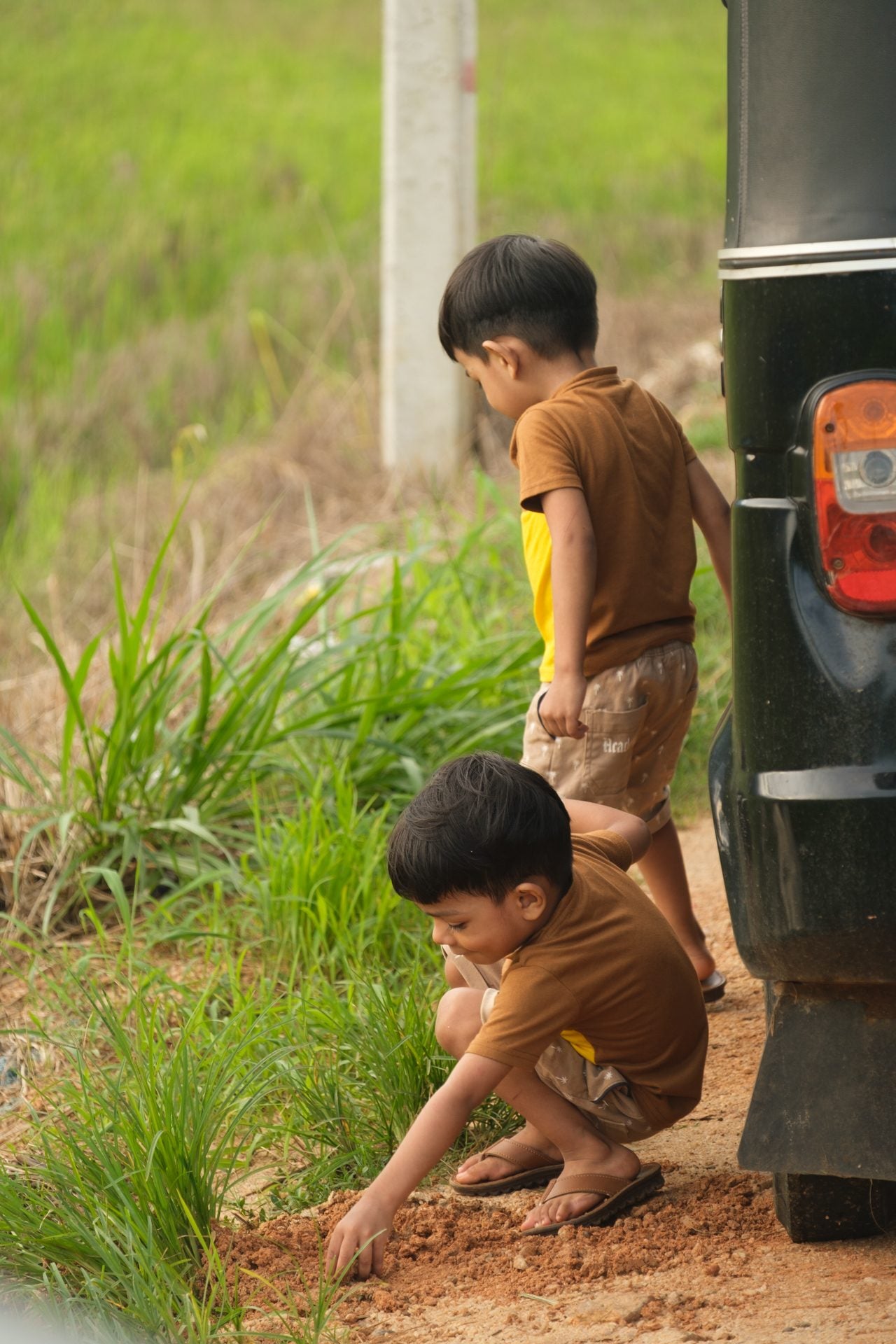 Two children crouching in the dirt beside a parked vehicle at the field edge