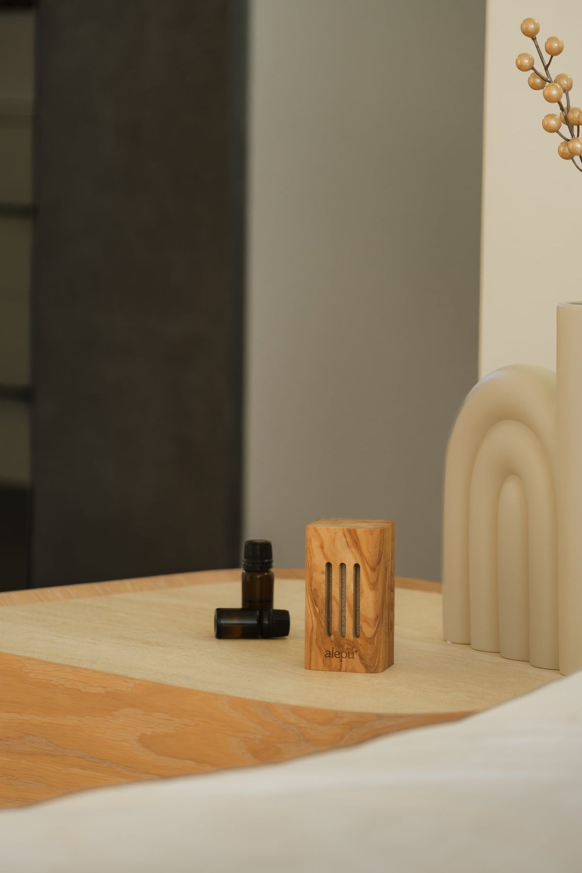 The square olive wood diffuser with two small oil bottles on a bedside table