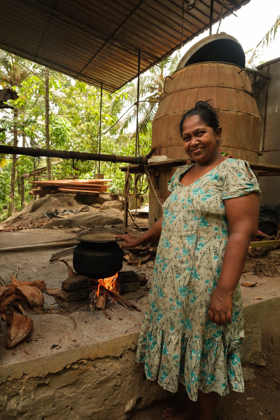 A distiller standing beside a wood-fired still under a tin roof in Sri Lanka, smiling