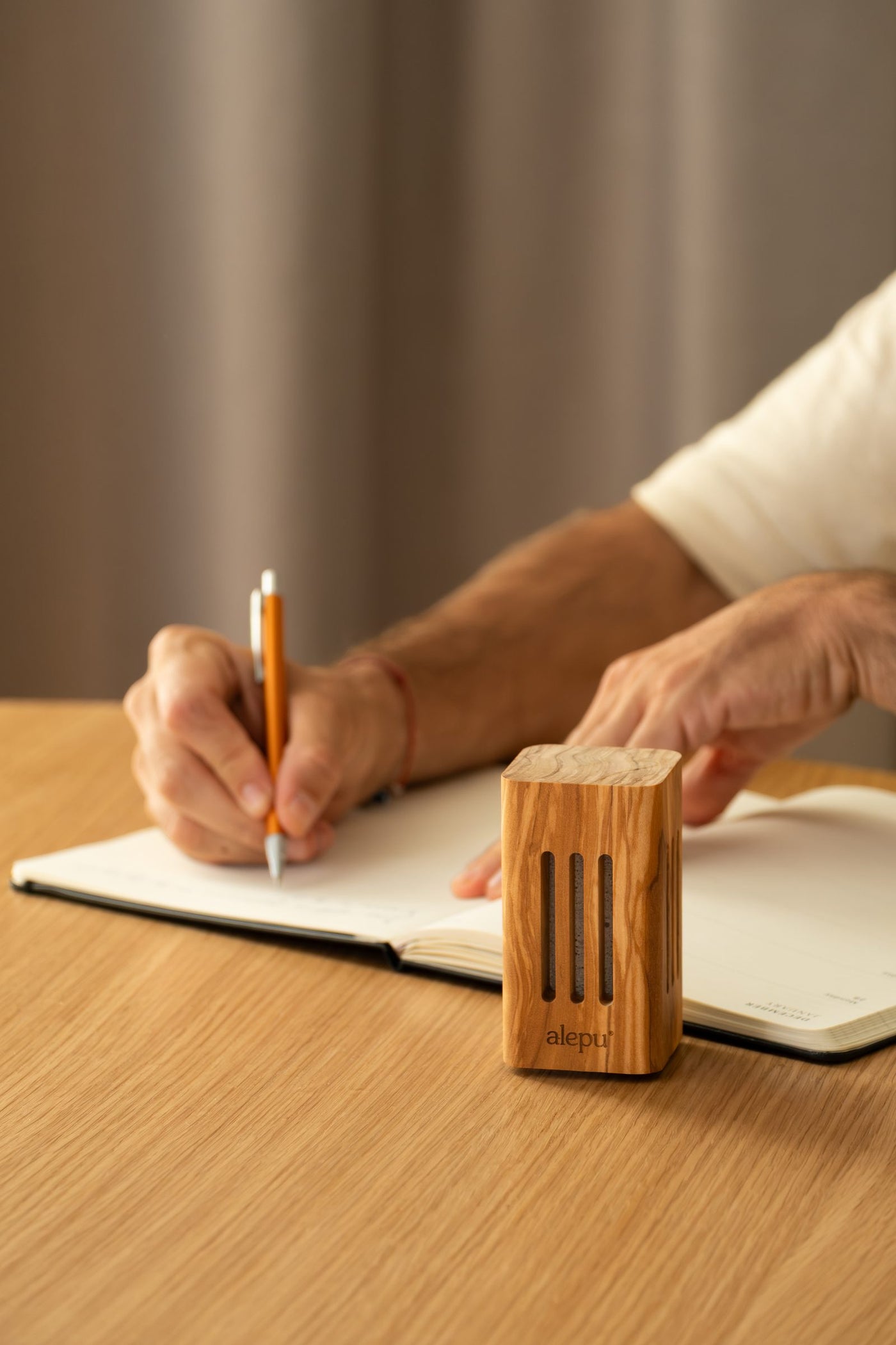 A square olive wood diffuser on a table while someone writes in a notebook