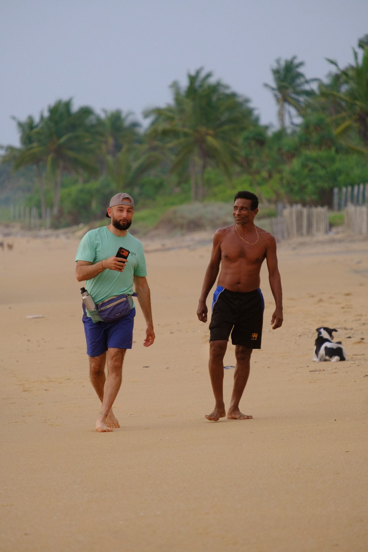 Two men walking together along an empty beach at dusk, palms behind them