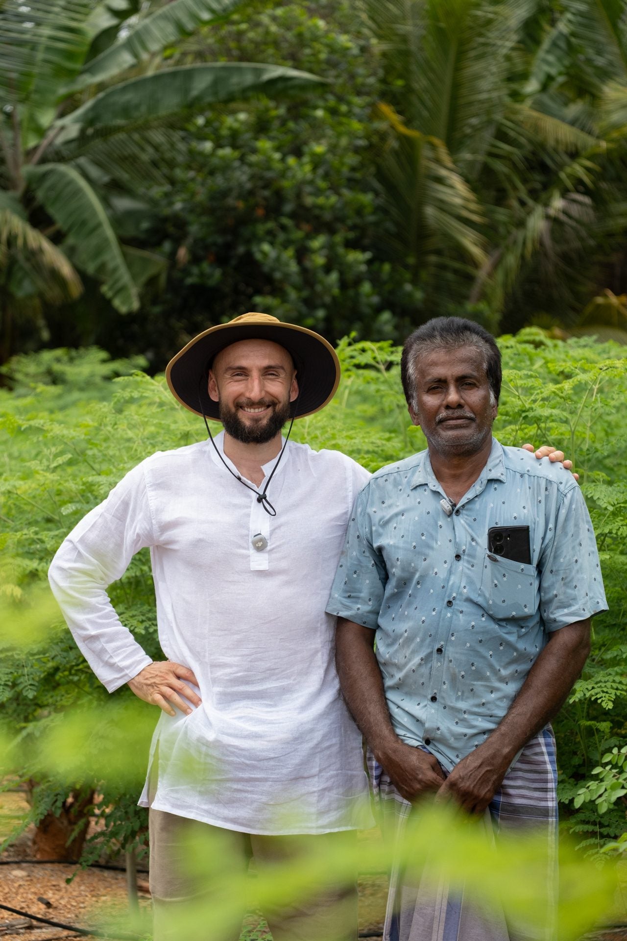 Two men standing together at the edge of a planted field, one in a wide-brimmed hat